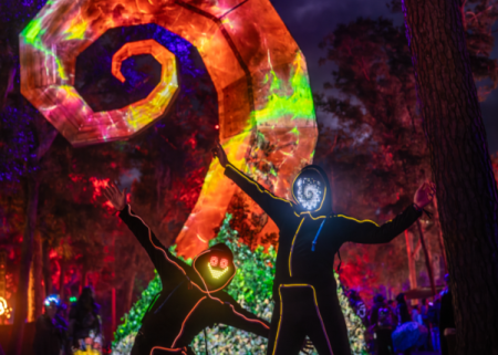 Two performers pose before a glowing spiral installation in the forest