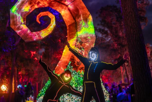 Two performers pose before a glowing spiral installation in the forest